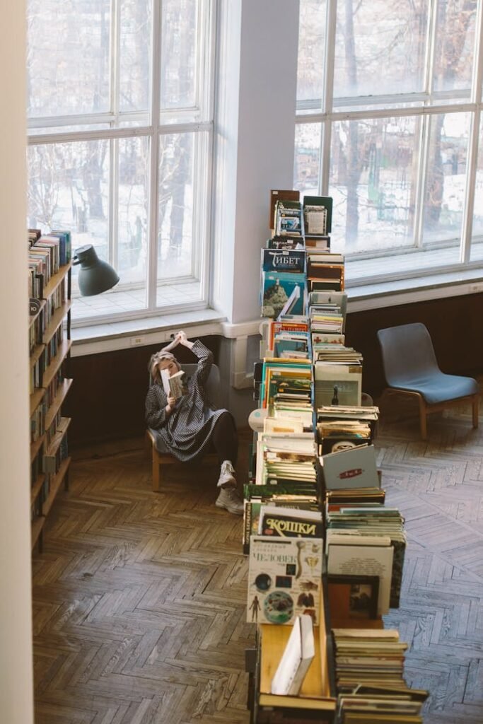 A quiet library corner with a person reading, surrounded by wooden bookshelves and large windows.